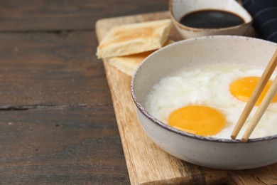 Photo of Traditional asian breakfast with half-boiled eggs in bowl, soy sauce, bread and chopsticks on wooden table, closeup. Space for text