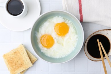 Photo of Traditional asian breakfast with half-boiled eggs in bowl, soy sauce, bread, chopsticks and coffee on white tiled table, flat lay