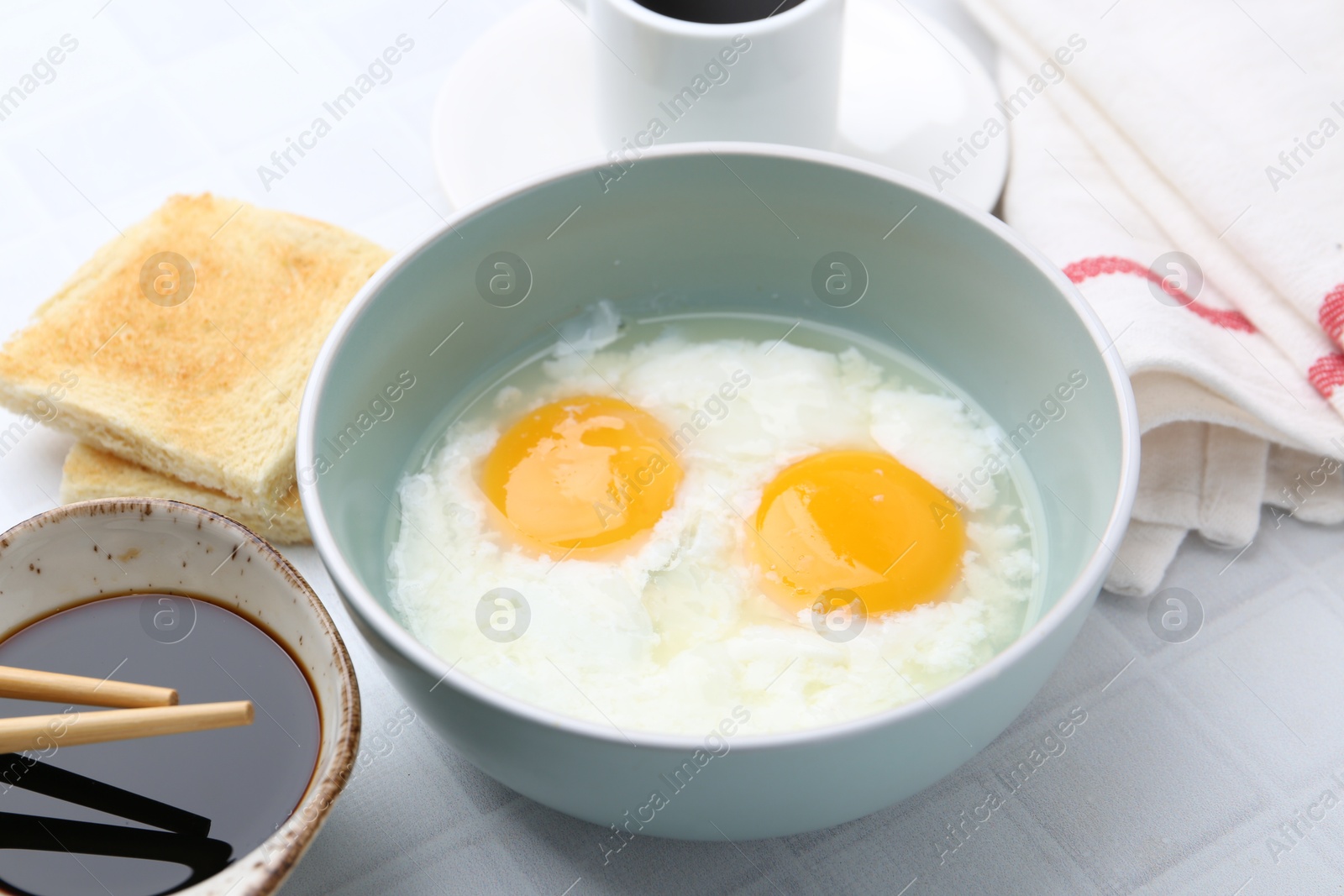 Traditional asian breakfast with half-boiled eggs in bowl, soy sauce, bread, chopsticks and coffee on white tiled table, closeup Photo of Traditional asian breakfast with half-boiled eggs in bowl, soy sauce, bread, chopsticks and coffee on white tiled table, closeup