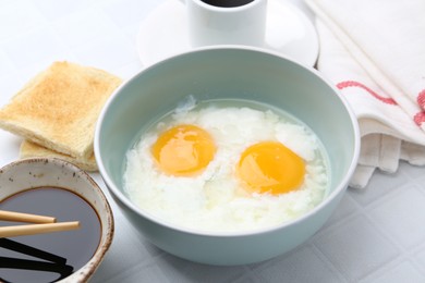Photo of Traditional asian breakfast with half-boiled eggs in bowl, soy sauce, bread, chopsticks and coffee on white tiled table, closeup