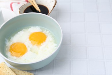 Photo of Traditional asian breakfast with half-boiled eggs in bowl, soy sauce, bread and chopsticks on white tiled table, closeup. Space for text