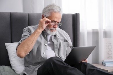 Photo of Senior man with vision problem using laptop on bed at home