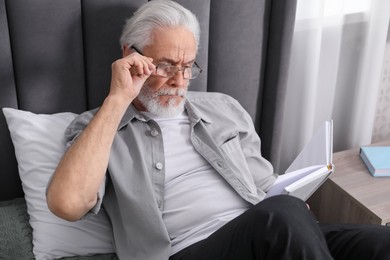 Photo of Senior man with vision problem reading book on bed at home