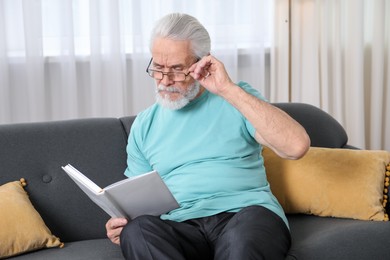 Photo of Senior man with vision problem reading book on sofa at home