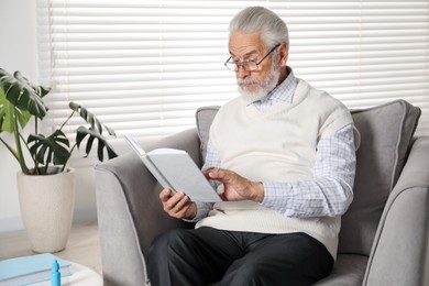 Photo of Senior man with vision problem reading book in armchair at home