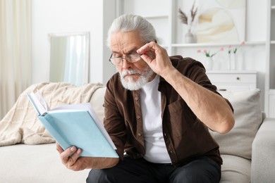 Photo of Senior man with vision problem reading book on sofa at home