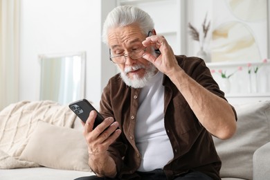 Photo of Senior man with vision problem using smartphone on sofa at home, space for text