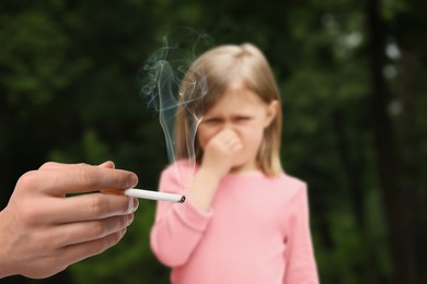 Image of Passive smoking. Little girl covering her nose with hand because of man smoking nearby outdoors. Selective focus