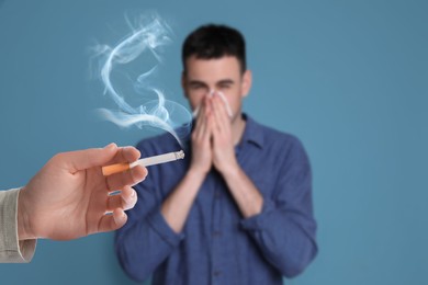 Image of Passive smoking. Man covering his nose with tissue because of woman smoking nearby on light blue background. Selective focus