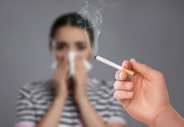Image of Passive smoking. Woman covering her nose with tissue because of man smoking nearby on grey background. Selective focus