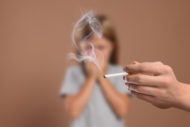 Image of Passive smoking. Little girl covering her nose with hands because of man smoking nearby on pale brown background. Selective focus
