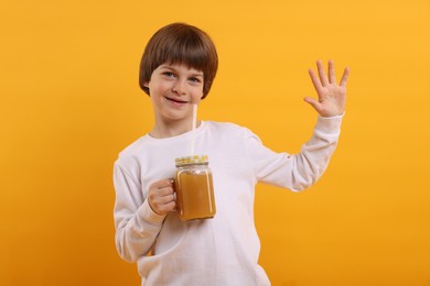 Photo of Cute little boy with mason jar of juice waving hello on orange background. Refreshing drink