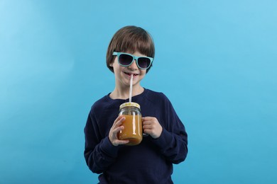 Photo of Cute little boy in sunglasses drinking orange juice on light blue background. Refreshing drink