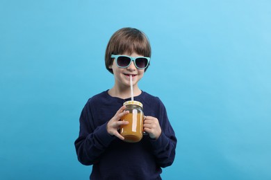 Photo of Cute little boy in sunglasses drinking orange juice on light blue background. Refreshing drink