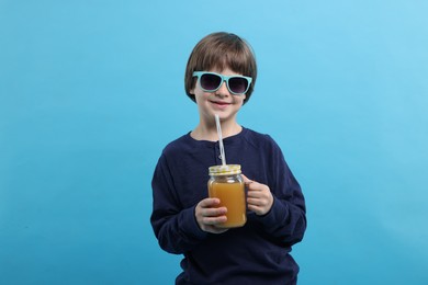 Photo of Happy little boy in sunglasses with mason jar of orange juice on light blue background. Refreshing drink