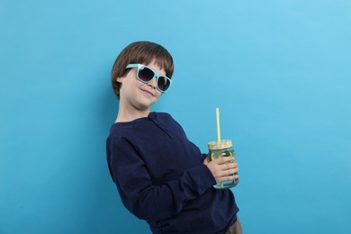 Photo of Cute little boy in sunglasses with mason jar of lemonade on light blue background. Refreshing drink