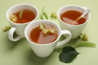 Photo of Tasty linden tea in cups, spoon, leaves and flowers on light green background, closeup