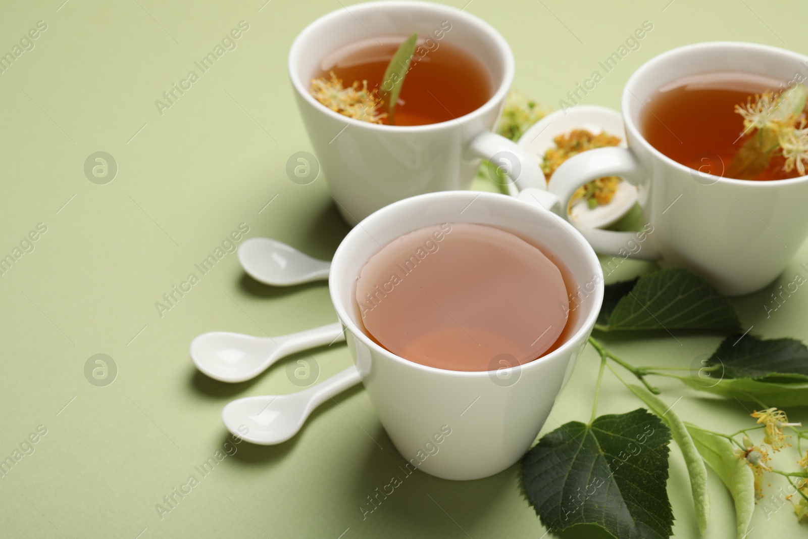 Tasty linden tea in cups, spoons, leaves and flowers on light green background, closeup. Space for text Photo of Tasty linden tea in cups, spoons, leaves and flowers on light green background, closeup. Space for text