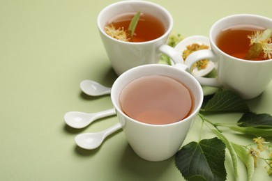 Photo of Tasty linden tea in cups, spoons, leaves and flowers on light green background, closeup. Space for text