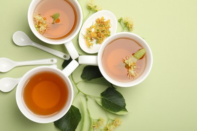 Photo of Tasty linden tea in cups, spoons, leaves and flowers on light green background, flat lay. Space for text