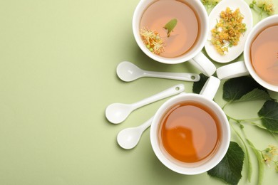 Photo of Tasty linden tea in cups, spoons, leaves and flowers on light green background, flat lay. Space for text