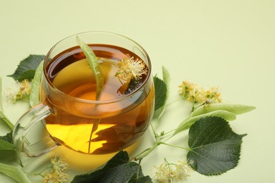 Photo of Tasty linden tea in cup, leaves and flowers on light green background, closeup