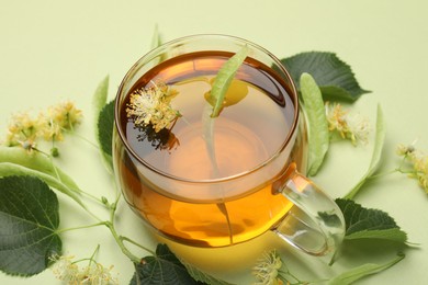 Photo of Tasty linden tea in cup, leaves and flowers on light green background, closeup