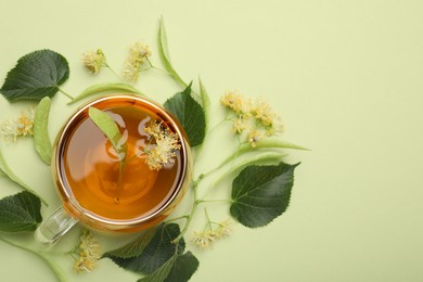 Photo of Tasty linden tea in cup, leaves and flowers on light green background, flat lay. Space for text