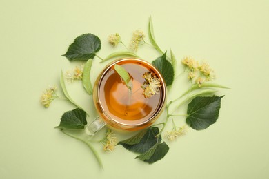 Photo of Tasty linden tea in cup, leaves and flowers on light green background, flat lay