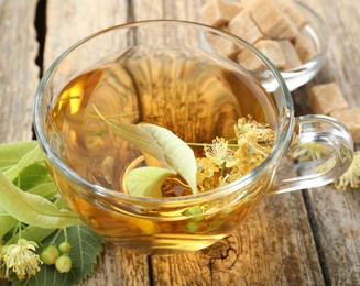 Photo of Tasty linden tea in cup, sugar cubes, leaves and flowers on wooden table, closeup
