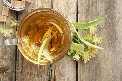 Photo of Tasty linden tea in cup, sugar cubes, leaves and flowers on wooden table, flat lay