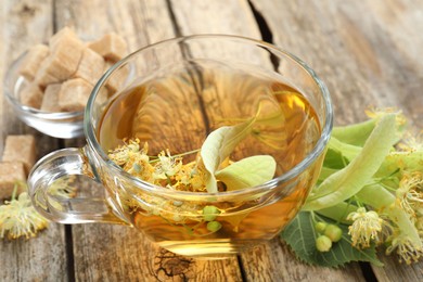 Photo of Tasty linden tea in cup, sugar cubes, leaves and flowers on wooden table, closeup