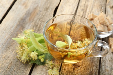 Photo of Tasty linden tea in cup, sugar cubes, leaves and flowers on wooden table, closeup