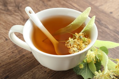 Photo of Tasty linden tea in cup, leaves and flowers on wooden table, closeup