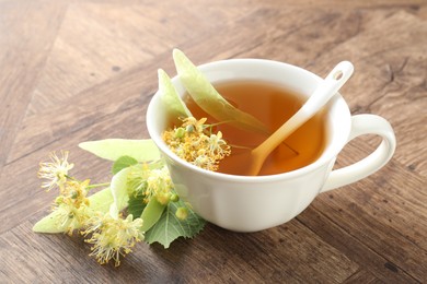 Photo of Tasty linden tea in cup, leaves and flowers on wooden table, closeup