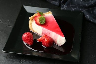 Photo of Piece of tasty cheesecake with strawberries, sauce and mint on black table, closeup