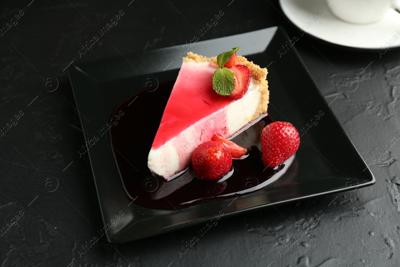Photo of Piece of tasty cheesecake with strawberries, sauce and mint on black table, closeup