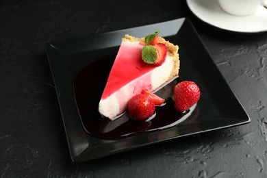 Photo of Piece of tasty cheesecake with strawberries, sauce and mint on black table, closeup