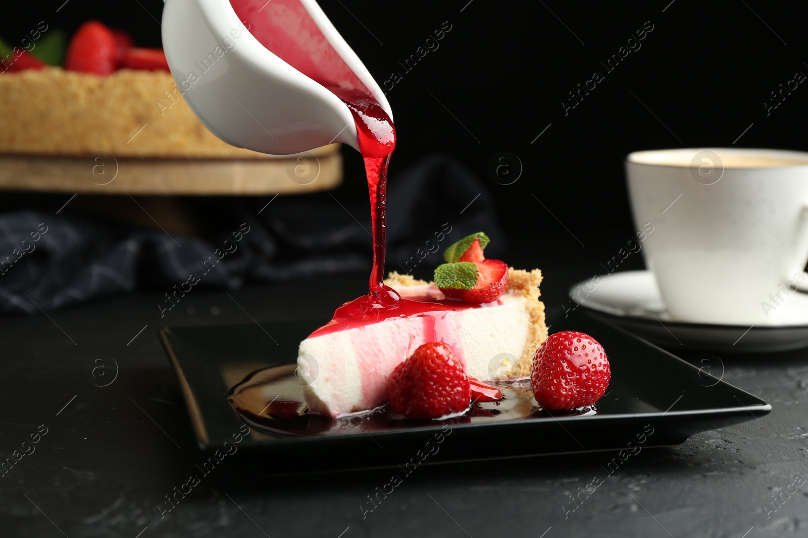 Photo of Pouring sauce onto piece of tasty cheesecake with strawberries and mint at black table, closeup