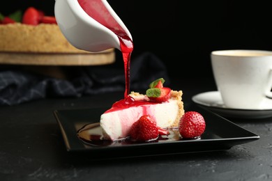 Photo of Pouring sauce onto piece of tasty cheesecake with strawberries and mint at black table, closeup