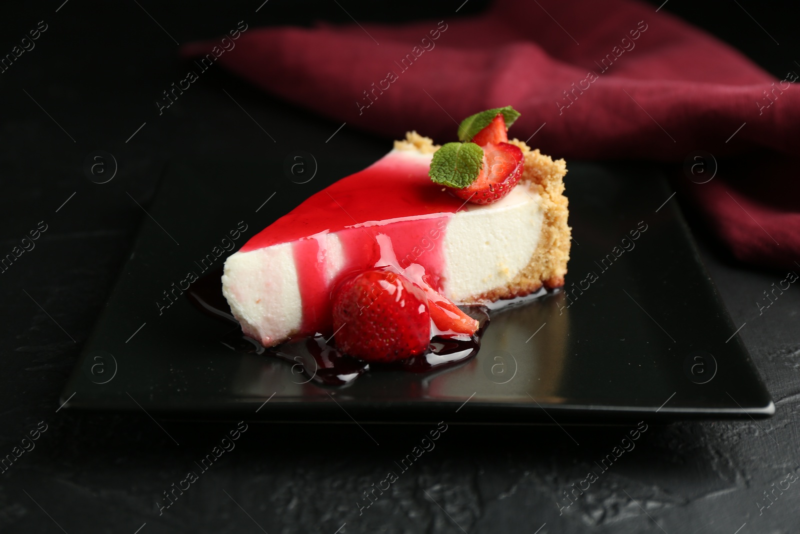 Photo of Piece of tasty cheesecake with strawberries, sauce and mint on black table, closeup