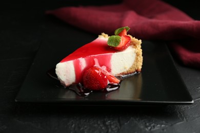 Photo of Piece of tasty cheesecake with strawberries, sauce and mint on black table, closeup