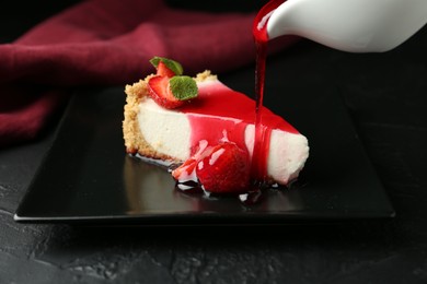 Photo of Pouring sauce onto piece of tasty cheesecake with strawberries and mint at black table, closeup