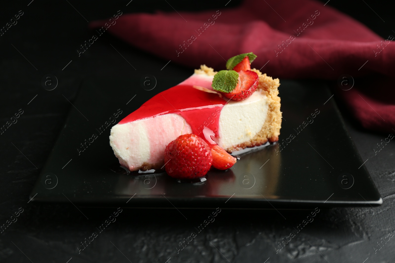 Photo of Piece of tasty cheesecake with strawberries, sauce and mint on black table, closeup