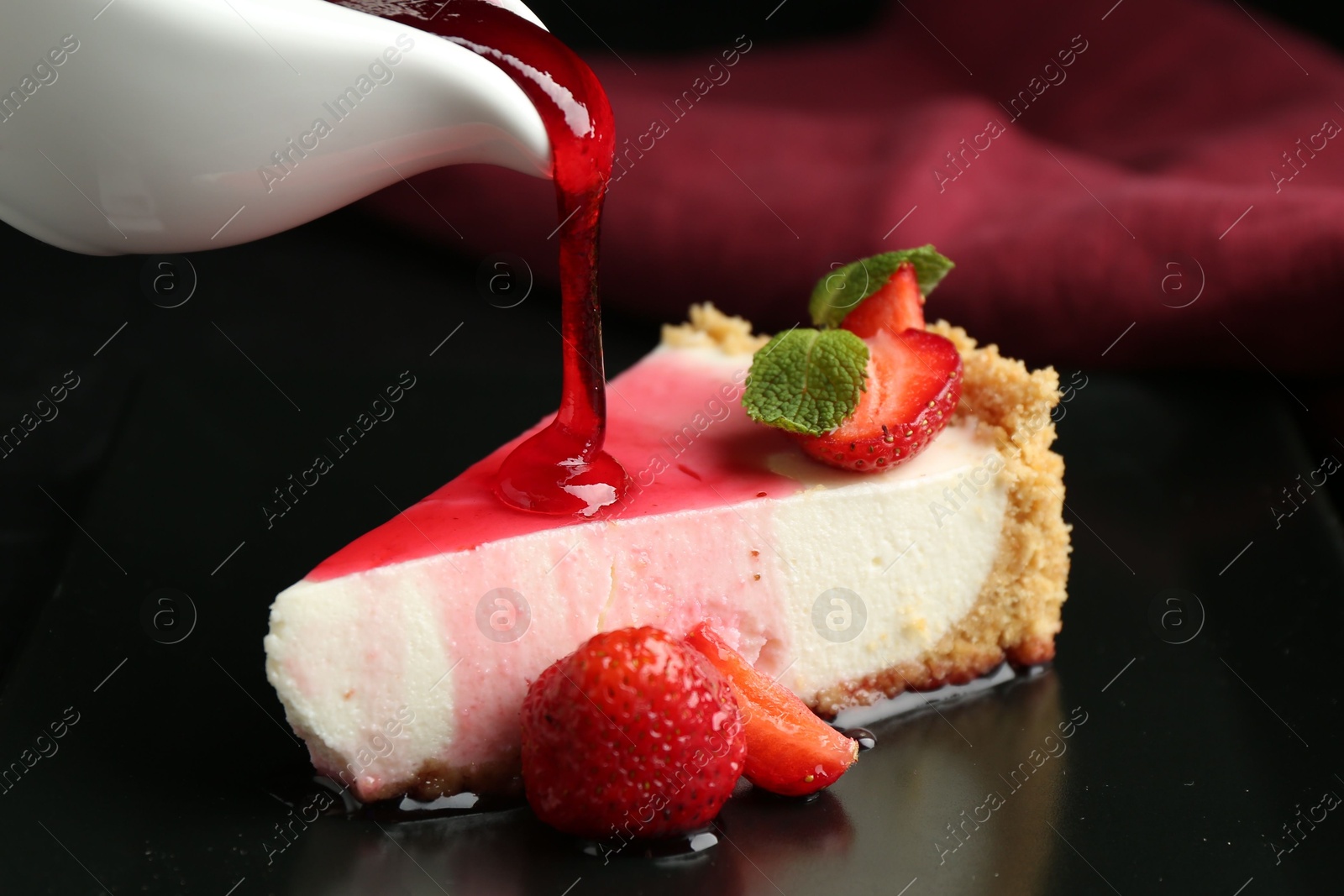 Photo of Pouring sauce onto piece of tasty cheesecake with strawberries and mint at table, closeup