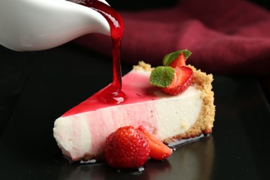 Photo of Pouring sauce onto piece of tasty cheesecake with strawberries and mint at table, closeup