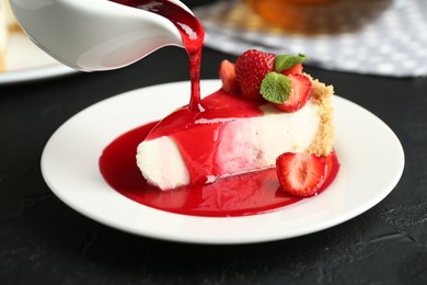 Photo of Pouring sauce onto piece of tasty cheesecake with strawberries and mint at black table, closeup