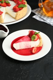 Photo of Piece of tasty cheesecake with strawberries, sauce and mint on black table, closeup