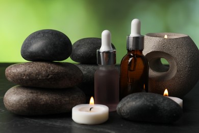 Photo of Spa composition with stones, droppers and burning candles on dark table against blurred green background, closeup