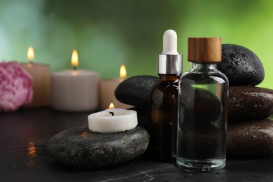 Photo of Spa composition with wet stones, cosmetic products and burning candles on dark textured table against blurred green background, selective focus